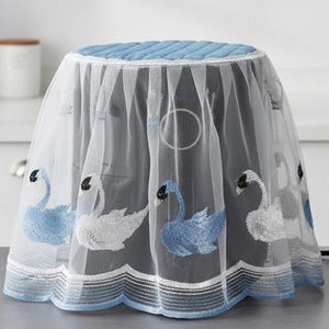 Può includere: Una copertura bianca e trasparente per elettrodomestici con bordo smerlato blu e cigni ricamati. La copertura ha una parte superiore trapuntata blu chiaro. A destra c'è una piccola pianta in vaso.