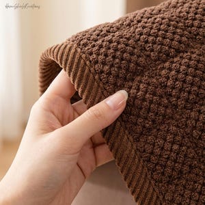 May include: Close-up of a brown textured fabric with a raised, dotted pattern. The edge of the fabric has a ribbed border. The fabric is being held by a hand. The HomeShieldCreations logo is in the top left corner.