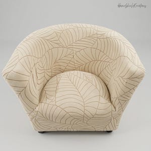 Könnte beinhalten: Ein beigefarbener Sessel mit einem Blattmuster in dunklerem Beige. Der Stuhl hat eine abgerundete Rückenlehne und Armlehnen sowie einen geschwungenen Sitz. Der Stuhl steht auf einem hellgrauen Hintergrund. Der Text "HomeShieldCreations" befindet sich in der oberen rechten Ecke.