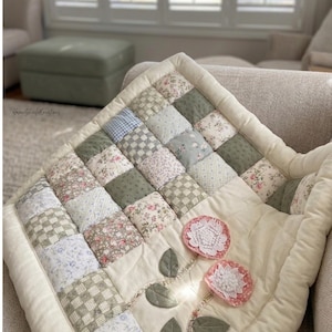Puede incluir: Un edredón de patchwork hecho a mano con un diseño floral. El edredón presenta cuadrados de varios patrones y colores, incluyendo estampados florales, cuadros y verdes y azules lisos. Dos flores de tela rosa con centros de encaje blanco están adjuntas.