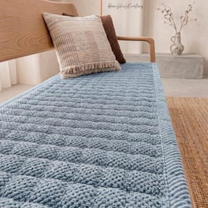 May include: A light blue textured blanket with a repeating pattern, placed on a wooden bench. Two decorative pillows with fringe and a brown pillow are on the bench. The blanket has a soft, plush appearance.
