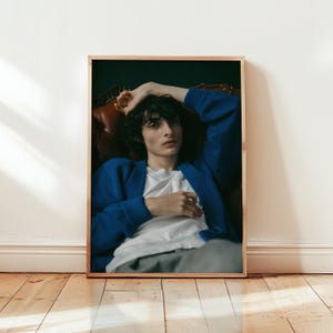 Op de afbeelding: Een ingelijste foto met een persoon die een blauw vest draagt over een wit t-shirt. De persoon zit op een bruine leren bank. De foto wordt weergegeven op een houten vloer tegen een witte muur.