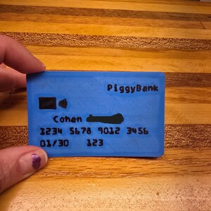 May include: A blue piggy bank shaped like a credit card. The card has the word "PiggyBank" printed on it, along with a name, numbers, and other details. The card is being held by a person.