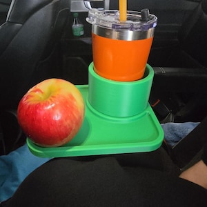 May include: A green car tray holds an orange drink in a stainless steel tumbler with a straw, and a red and yellow apple. The tray is placed on a person's lap inside a car. A small bottle is visible in the background.