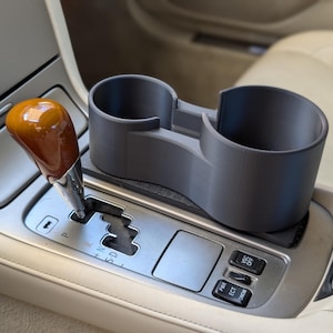 May include: A gray, 3D-printed car cup holder with two circular compartments. The cup holder sits on the center console of a vehicle, next to the gear shift. The gear shift has a wooden knob.