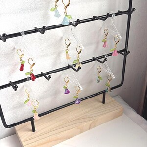 May include: A selection of flower earrings with gold-coloured hooks. The earrings are in various colours, including pink, red, purple, and blue, with green leaf accents. The earrings are displayed on a black metal rack with a wooden base.