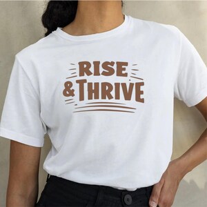 以下が含まれることがあります： 「RISE & THRIVE」の茶色の文字がプリントされた白いTシャツ。デザインには、テキストの上と下に水平線があり、シンプルながらもインパクトのあるグラフィックを作成しています。