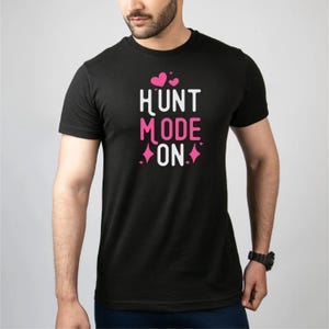以下が含まれることがあります： 白とピンクで「HUNT MODE ON」の文字が入った黒いTシャツ。デザインにはピンクのハートとひし形が含まれています。カジュアルな服装です。