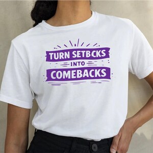 Könnte beinhalten: Weißes T-Shirt mit dem lila Text "TURN SETBCKS INTO COMEBACKS". Der Text ist in zwei Zeilen mit einem dekorativen Burst darüber angeordnet. Ein einfaches, lässiges Oberteil für den Alltag.