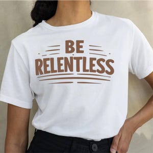 Peut inclure: T-shirt blanc avec l'inscription marron "BE RELENTLESS". Le texte est centré et souligné de traits horizontaux. Le t-shirt a des manches courtes et un col rond.