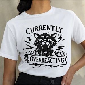 May include: White t-shirt featuring a black graphic of an angry cat with the words "CURRENTLY OVERREACTING". The cat graphic includes lightning bolts and a banner.