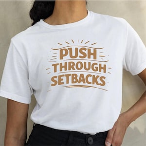 May include: White t-shirt with the phrase "PUSH THROUGH SETBACKS" in brown lettering. The design includes stylized lines and sunburst elements. The shirt is made of a soft material.