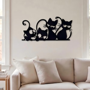 May include: A black metal wall art piece featuring a silhouette design of four stylized cats standing in a row. The cats have large eyes, pointed ears, and curved tails, connected by a single horizontal base line mounted on a plain wall.