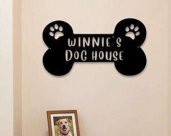 May include: A black wall-mounted sign shaped like a dog bone with paw print cutouts on each end. The text "WINNIE'S DOG HOUSE" is printed in white, sans-serif capital letters across the center of the bone-shaped sign.