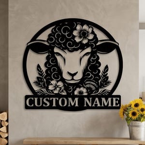 May include: Black metal wall art featuring a sheep's head with floral accents and the text "CUSTOM NAME" on a rectangular plaque. The sheep is set within a circular frame. Decorative home decor.
