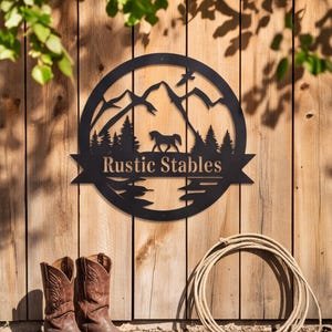 May include: A circular black metal wall sign featuring a cutout design of mountains, pine trees, a horse, and a bird. A horizontal banner across the center displays the text "Rustic Stables" in a serif font. The sign is mounted on a wooden fence.