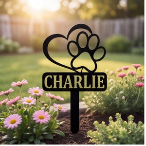 May include: A black metal garden stake featuring a cutout heart and paw print design above a rectangular plaque with the name CHARLIE in gold lettering. The stake is positioned vertically in dark soil among pink flowers and green foliage.