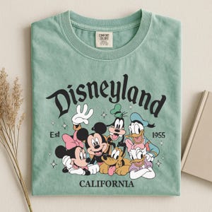 K&ouml;nnte beinhalten: Salbeigr&uuml;nes T-Shirt mit einem Aufdruck von Disney-Figuren, darunter Micky und Minnie Maus, Donald Duck und Goofy. Das Wort "Disneyland" ist oben gew&ouml;lbt, mit "California" unten. Das T-Shirt ist mit "Comfort Colors" gekennzeichnet.