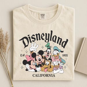 K&ouml;nnte beinhalten: Cremefarbenes T-Shirt mit einer Grafik von Disneyland-Charakteren, darunter Mickey und Minnie Mouse, Goofy, Donald und Daisy Duck sowie Pluto. Das Wort "Disneyland" steht &uuml;ber den Figuren, darunter "California". Das Shirt hat auch den Text "Est 1955".