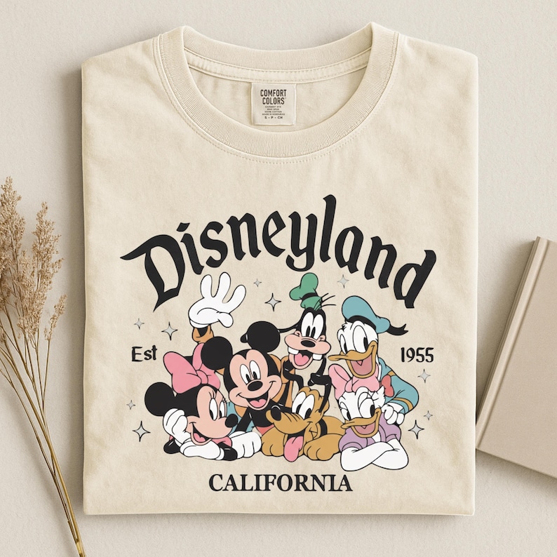 K&ouml;nnte beinhalten: Cremefarbenes T-Shirt mit einer Grafik von Disneyland-Charakteren, darunter Mickey und Minnie Mouse, Goofy, Donald und Daisy Duck sowie Pluto. Das Wort "Disneyland" steht &uuml;ber den Figuren, darunter "California". Das Shirt hat auch den Text "Est 1955".