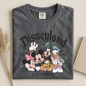 K&ouml;nnte beinhalten: Dunkelgraues T-Shirt mit einer Grafik von Disneyland-Charakteren, darunter Micky und Minnie Maus, Goofy, Donald und Daisy Duck und Pluto. Das Wort "Disneyland" ist oben gew&ouml;lbt, mit "California" unten und "Est 1955".