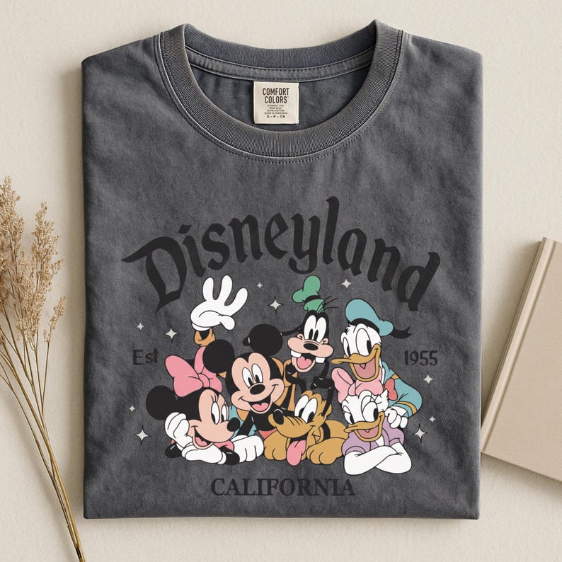 K&ouml;nnte beinhalten: Dunkelgraues T-Shirt mit einer Grafik von Disneyland-Charakteren, darunter Micky und Minnie Maus, Goofy, Donald und Daisy Duck und Pluto. Das Wort "Disneyland" ist oben gew&ouml;lbt, mit "California" unten und "Est 1955".