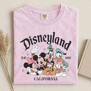 K&ouml;nnte beinhalten: Hellrosa T-Shirt mit einem Aufdruck von Disney-Figuren, darunter Micky Maus, Minnie Maus, Goofy, Donald Duck und Pluto. Das Wort "Disneyland" ist &uuml;ber den Figuren gew&ouml;lbt, darunter "California". Das T-Shirt ist gefaltet.