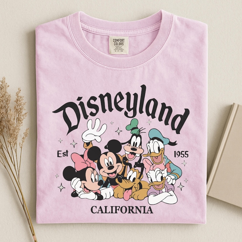 K&ouml;nnte beinhalten: Hellrosa T-Shirt mit einem Aufdruck von Disney-Figuren, darunter Micky Maus, Minnie Maus, Goofy, Donald Duck und Pluto. Das Wort "Disneyland" ist &uuml;ber den Figuren gew&ouml;lbt, darunter "California". Das T-Shirt ist gefaltet.