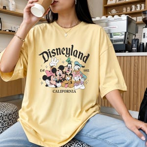 K&ouml;nnte beinhalten: Hellgelbes T-Shirt mit dem Disneyland-Logo und den Zeichentrickfiguren Mickey, Minnie, Goofy, Donald und Daisy Duck. Das Shirt hat kurze &Auml;rmel und den Text "Est 1955 CALIFORNIA".