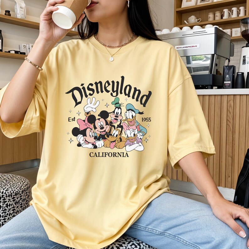 K&ouml;nnte beinhalten: Hellgelbes T-Shirt mit dem Disneyland-Logo und den Zeichentrickfiguren Mickey, Minnie, Goofy, Donald und Daisy Duck. Das Shirt hat kurze &Auml;rmel und den Text "Est 1955 CALIFORNIA".
