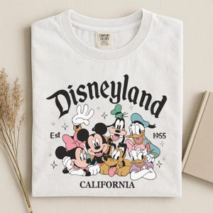 K&ouml;nnte beinhalten: Wei&szlig;es T-Shirt mit einem Aufdruck von Disneyland-Charakteren, darunter Micky und Minnie Maus, Goofy, Donald und Daisy Duck. Der Text "Disneyland California Est 1955" ist &uuml;ber den Figuren aufgedruckt.