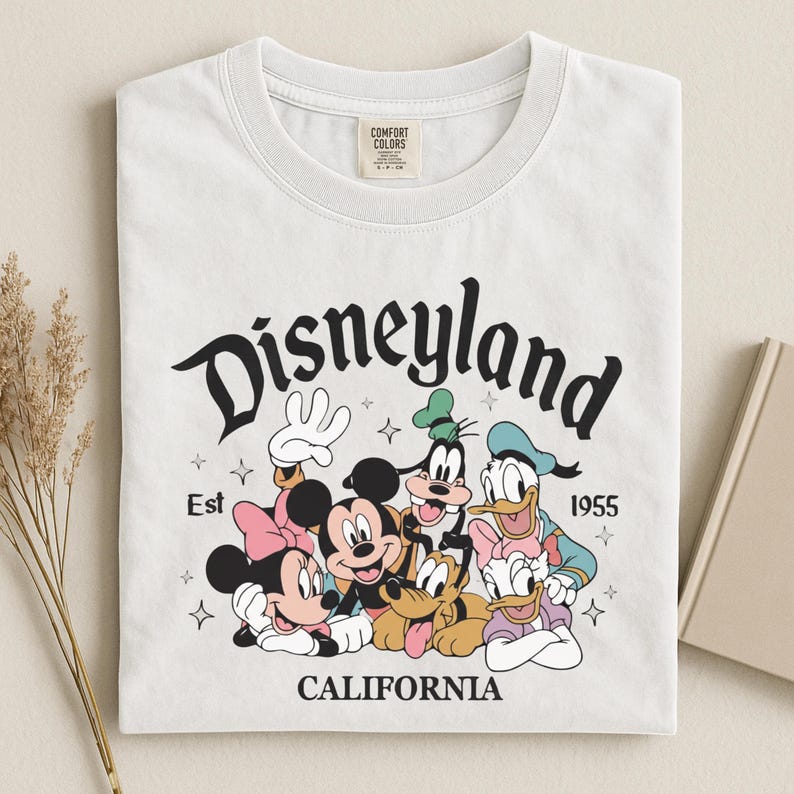 K&ouml;nnte beinhalten: Wei&szlig;es T-Shirt mit einem Aufdruck von Disneyland-Charakteren, darunter Micky und Minnie Maus, Goofy, Donald und Daisy Duck. Der Text "Disneyland California Est 1955" ist &uuml;ber den Figuren aufgedruckt.