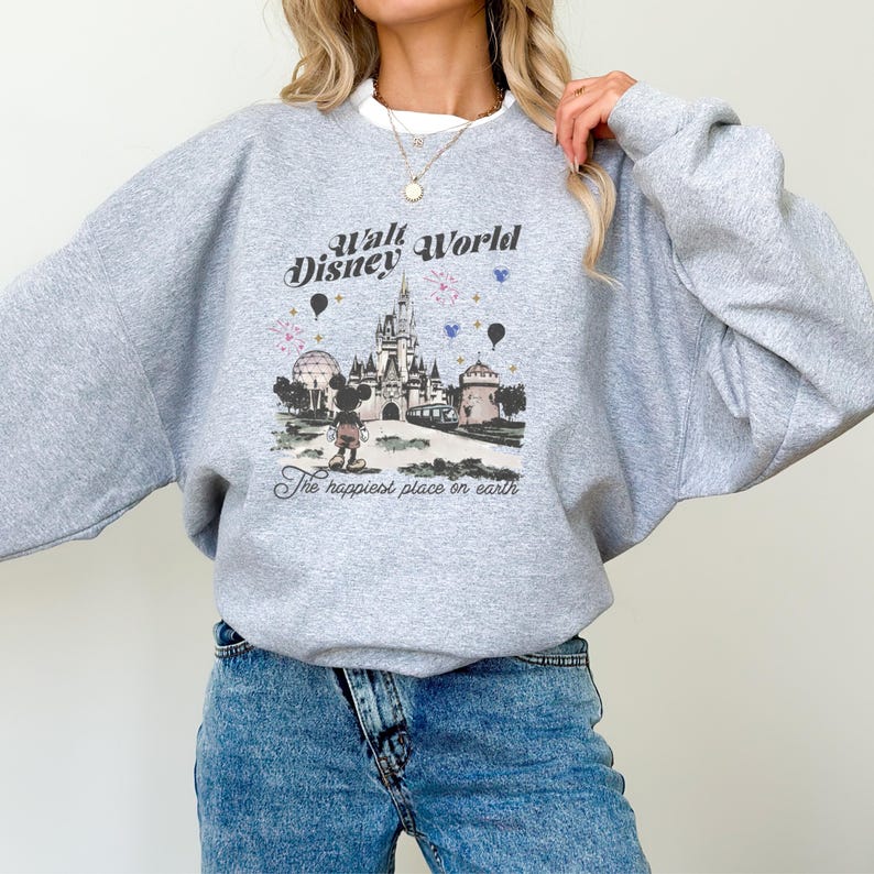 May include: A light gray sweatshirt featuring a graphic of Cinderella's castle, Mickey Mouse, and the text "Walt Disney World" and "The happiest place on earth." The design includes balloons and floral accents.