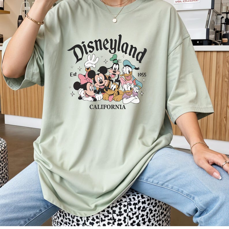 K&ouml;nnte beinhalten: Salbeigr&uuml;nes T-Shirt mit dem Disneyland-Logo und einer Grafik von Mickey Mouse, Minnie Mouse, Donald Duck, Goofy und Pluto. Der Text "Est 1955 California" ist ebenfalls aufgedruckt. Das T-Shirt hat kurze &Auml;rmel und eine lockere Passform.