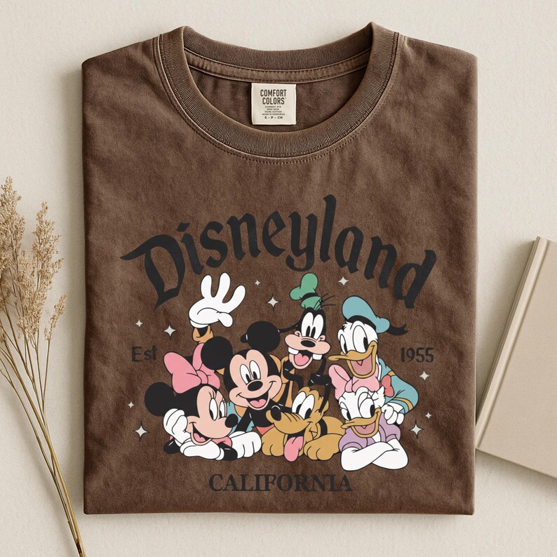 K&ouml;nnte beinhalten: Braunes T-Shirt mit dem Wort "Disneyland" in einer Vintage-Schriftart und einer Grafik von Micky Maus, Minnie Maus, Goofy, Donald Duck, Daisy Duck und Pluto. Das Shirt hat auch den Text "California" und "Est 1955".