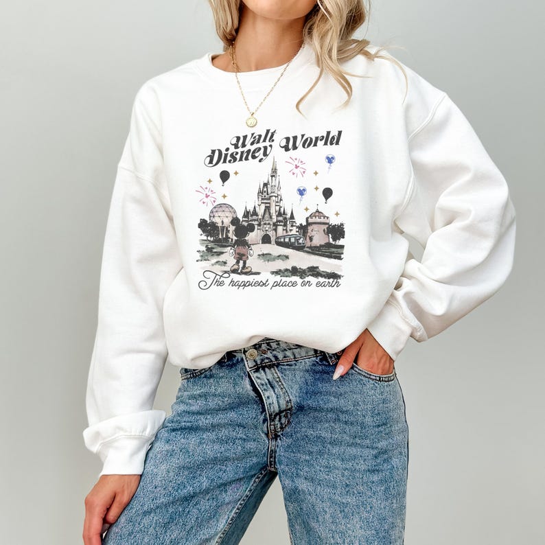 May include: White sweatshirt featuring a graphic of the Walt Disney World castle, fireworks, and Mickey Mouse. The text "Walt Disney World" and "The happiest place on earth" are also displayed. The sweatshirt is worn by a person wearing blue jeans.