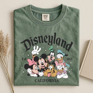 K&ouml;nnte beinhalten: Gr&uuml;nes T-Shirt mit dem Wort "Disneyland" in einer stilisierten Schriftart und dem Text "Est 1955 CALIFORNIA". Das Shirt zeigt die Zeichentrickfiguren Mickey Mouse, Minnie Mouse, Goofy, Donald Duck und Pluto.