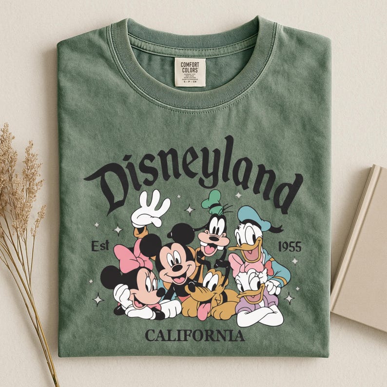 K&ouml;nnte beinhalten: Gr&uuml;nes T-Shirt mit dem Wort "Disneyland" in einer stilisierten Schriftart und dem Text "Est 1955 CALIFORNIA". Das Shirt zeigt die Zeichentrickfiguren Mickey Mouse, Minnie Mouse, Goofy, Donald Duck und Pluto.