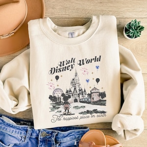 May include: Cream-colored sweatshirt featuring a graphic of Cinderella's castle, Mickey Mouse, and the text "Walt Disney World" and "The happiest place on earth." The design includes fireworks and balloons. The sweatshirt is paired with blue jeans and a hat.