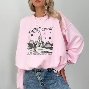 May include: A light pink sweatshirt featuring a graphic of Cinderella's castle, Mickey Mouse, and the text "Walt Disney World" and "The happiest place on earth." The sweatshirt has long sleeves.