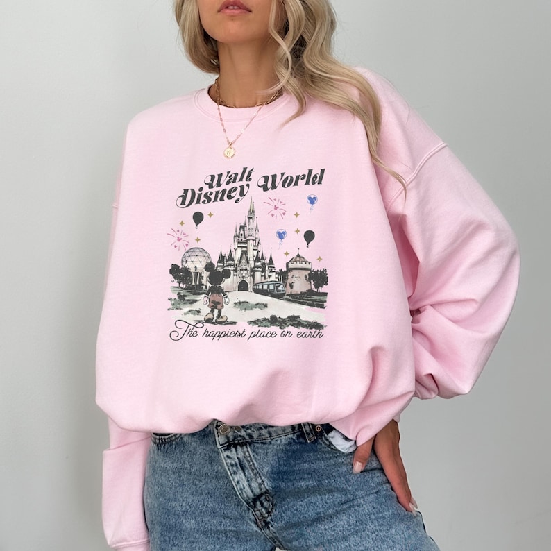 May include: A light pink sweatshirt featuring a graphic of Cinderella's castle, Mickey Mouse, and the text "Walt Disney World" and "The happiest place on earth." The sweatshirt has long sleeves.