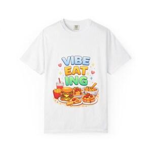 May include: White t-shirt featuring the text "Vibe Eat Ing" in a colourful design, with illustrations of fast food. The image includes a burger, fries, pizza, waffles, and a drink. The style of the design is cartoon-like.