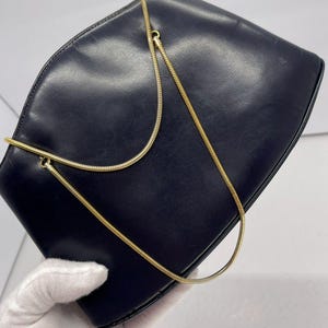 Puede incluir: Un clutch de cuero azul marino con una cadena dorada. El bolso tiene una parte superior curvada y un diseño simple y elegante. La cadena dorada es fina y delicada, añadiendo un toque de sofisticación al conjunto.