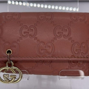 May include: A brown leather key case with an embossed pattern of interlocking "G" logos. The case has a gold-colored charm with the same logo. The key case is closed and appears to be new.