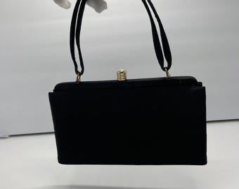 Puede incluir: Un bolso negro rectangular con un pequeño lazo en la parte superior de las asas. El bolso tiene un cierre dorado y asas negras. El bolso se sostiene sobre un fondo blanco.