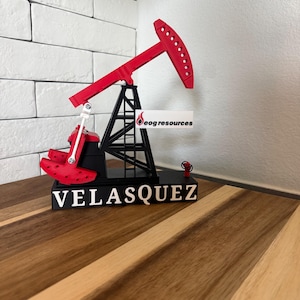 May include: A red and black oil pump jack model featuring a black base with the white text VELASQUEZ and a white sign attached to the structure displaying the EOG Resources logo. The mechanical pump jack has a red beam and a black support frame.