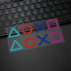 Puede incluir: Una pegatina transparente con los s&iacute;mbolos de los botones del mando de PlayStation: un tri&aacute;ngulo azul, un c&iacute;rculo rojo, una X azul y un cuadrado rosa. La pegatina est&aacute; sobre un teclado negro.