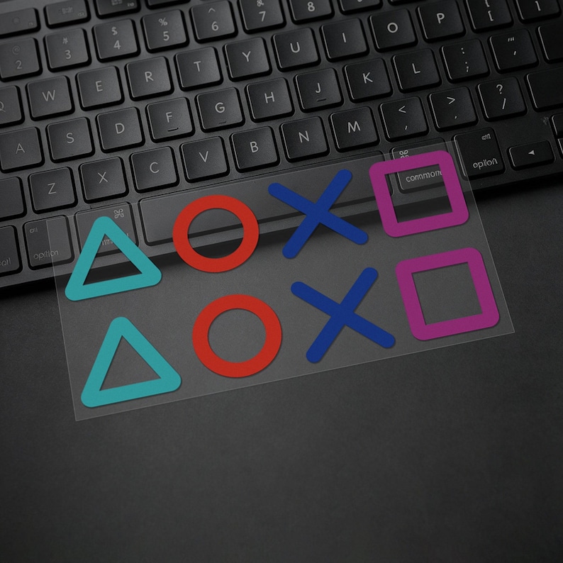Puede incluir: Una pegatina transparente con los s&iacute;mbolos de los botones del mando de PlayStation: un tri&aacute;ngulo azul, un c&iacute;rculo rojo, una X azul y un cuadrado rosa. La pegatina est&aacute; sobre un teclado negro.