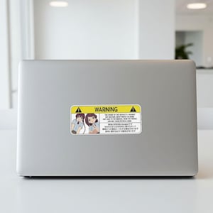 May include: A rectangular sticker with a yellow and black warning label. The label features anime-style illustrations of two girls and text in English and Japanese. The text warns about the vehicle owner's behavior.