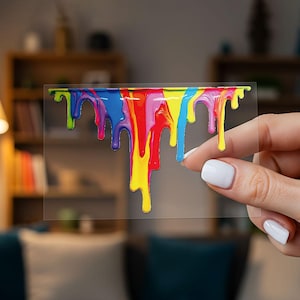 May include: A clear, rectangular sticker featuring a vibrant, dripping paint design. The paint drips in a rainbow of colors: green, blue, purple, red, yellow, and orange. The sticker is held by a person with white nail polish.